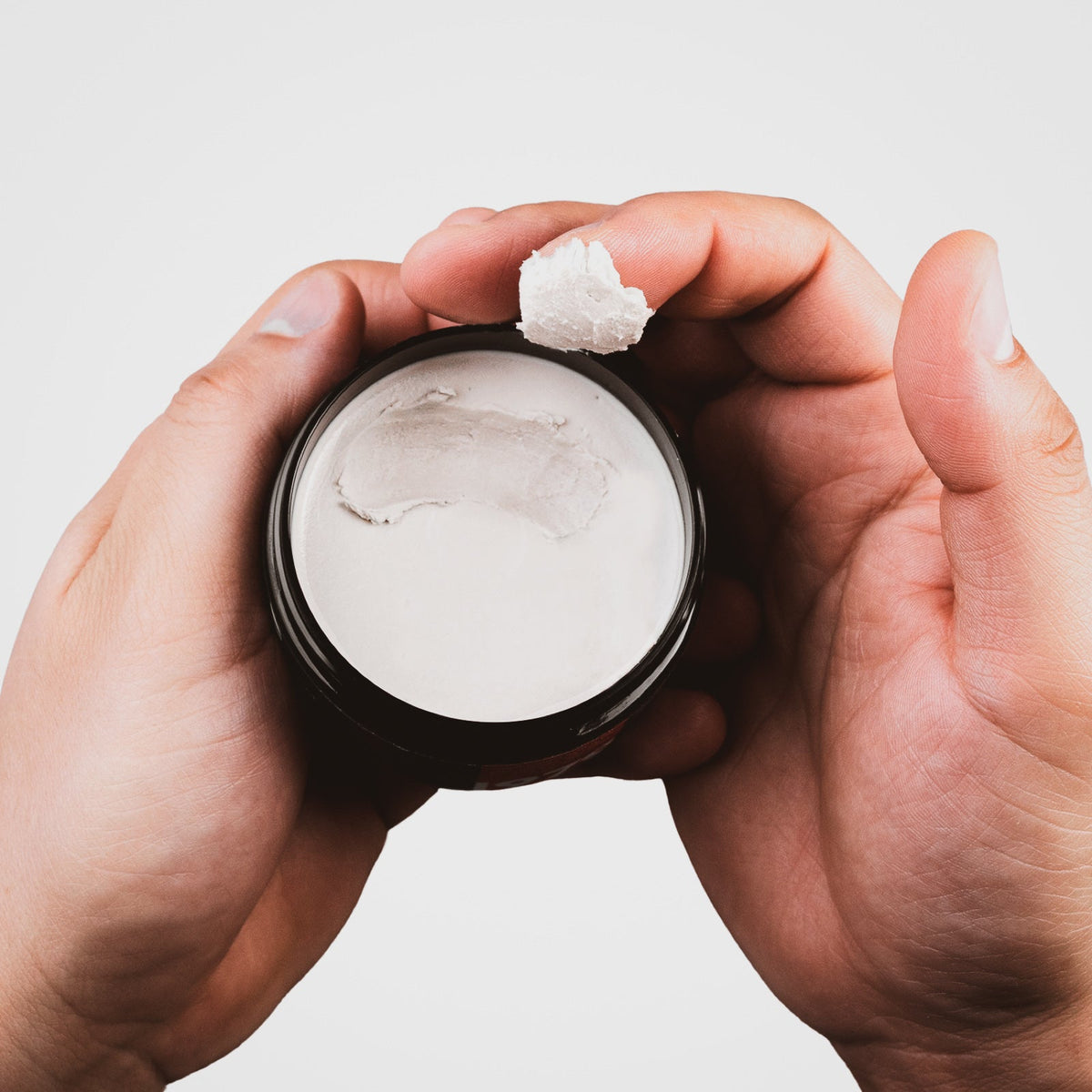 Hands holding an open jar of DASH FOR HIM Hair Clay, a scoop on the fingertip